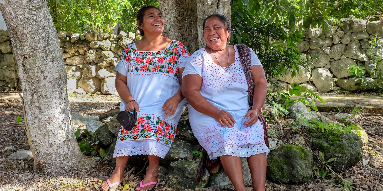 Two women sitting under a tree smiling.